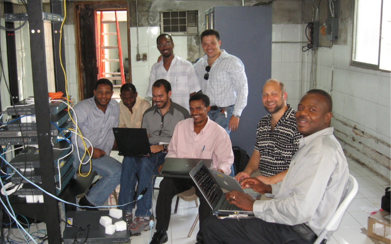 A group of men cluster around an Internet server, many typing on laptops.