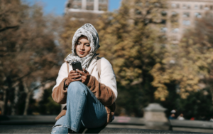 A woman wearing a headscarf and a jacket looks at her phone while sitting outside