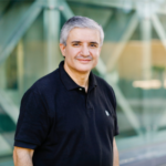 A man with short grey hair and a dark polo shirt smiles at the camera