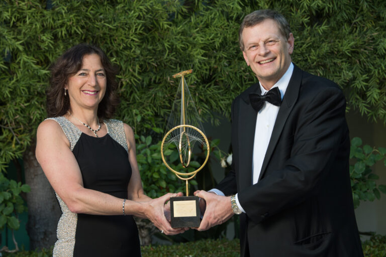 A woman wearing a dark gown and a man wearing a black tuxedo hold a gold award