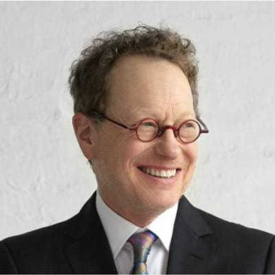 A man wearing a suit and round glasses smiles off to the side