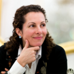 A woman with dark, curly hair wearing a button-down shirt and jacket looks off to the side with her chin on one hand