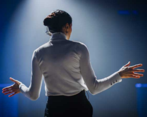 A woman wearing a turtleneck and dark pants stands on a stage, facing towards the audience with her arms held out as she speaks