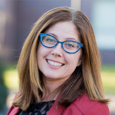 A woman with bright blue glasses and a colorful jacket smiles at the camera