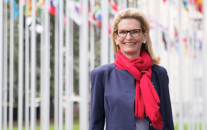 A woman wearing a bright scarf and jacket stands in front of a row of flags