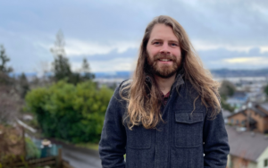 A smiling man with a beard and long hair standing in front of a cloudy sky and trees