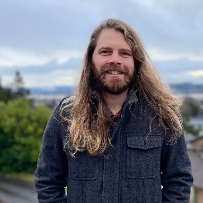 A smiling man with a beard and long hair standing in front of a cloudy sky and trees