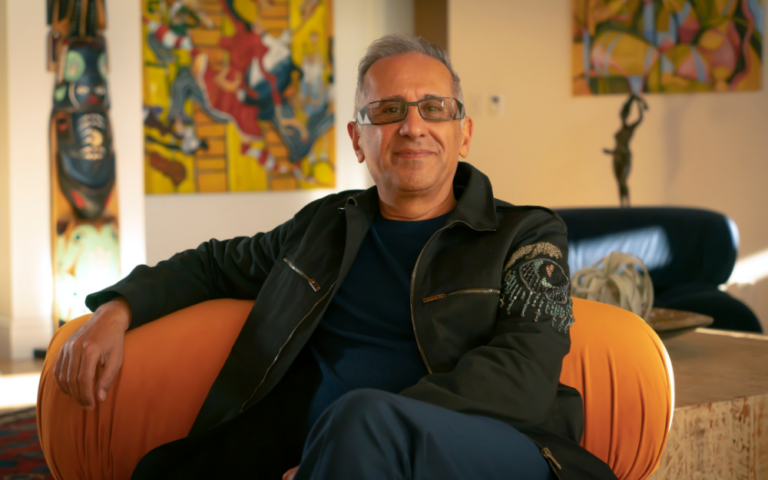 A man with glasses and an embroidered jacket lounges on an orange chair in front of colorful artwork