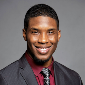 A man with short dark hair wearing a dark grey jacket and red dress shirt